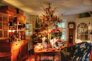 The cozy dining room holds more treasures, including a signature quilt (not seen) from Mark’s family, a redware-filled cupboard and a chandelier made by Denver tinsmith Charles Messner. In decorating the room for the holidays, Kerry utilized shades of red and pink for the floral arrangements that top the tables and are nestled in the chandelier. Note the pink-hued ornaments that fill a garden urn.