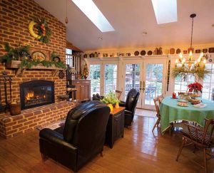 A fireplace is the focal point of the great room. Skylights and a wall of French doors admit plenty of natural light and provide a scenic view of the wooded property. The shelving over the doors holds Elaine's collection of antique crockery. A quilt covers the dining table.